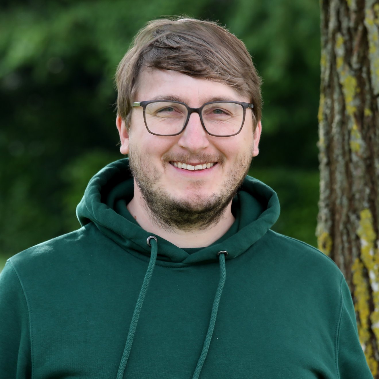 Ein lächelnder Mann mit Brille trägt einen grünen Hoodie. Er steht vor einem Baum und scheint in der Natur zu sein. Der Hintergrund ist verschwommen, sodass der Fokus auf dem Gesicht und der positiven Ausstrahlung des Mannes liegt.