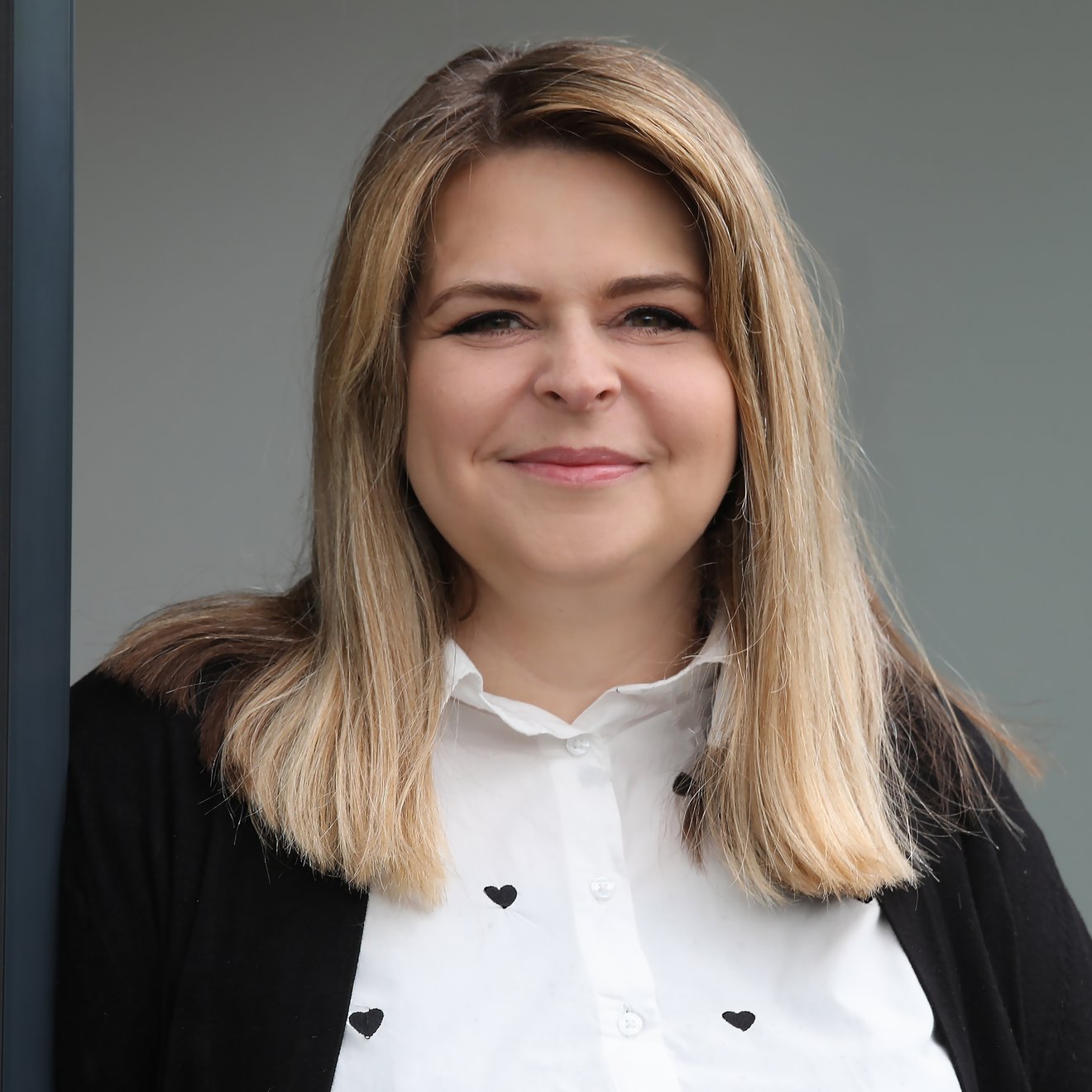 Eine lächelnde Frau mit langen, hellen Haaren steht vor einem neutralen Hintergrund. Sie trägt ein schwarzes Oberteil über einer weißen Bluse mit kleinen Herzen und wirkt freundlich und einladend.