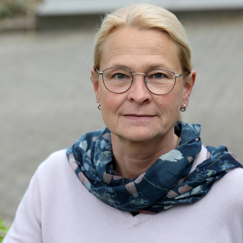 Porträt von Bianca Schmorl Foeste mit blonden, schulterlangen Haaren sie trägt eine Brille und einen bunten Schal. Sie blickt direkt in die Kamera und hat einen nachdenklichen Ausdruck.