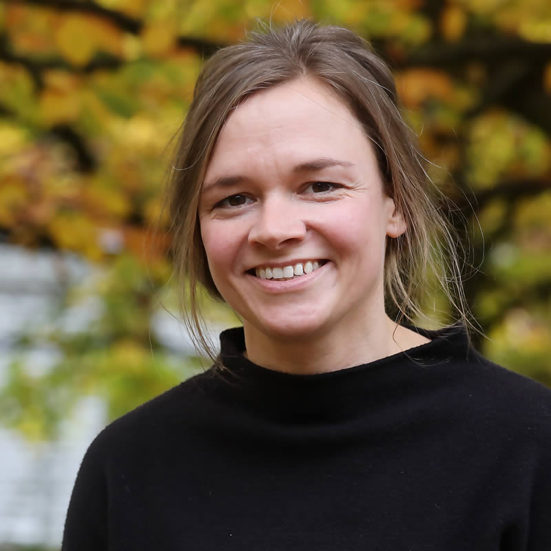 Portät von Julia Roemer mit schulterlangen, braunen Haaren sie trägt ein schwarzes Oberteil. Der Hintergrund zeigt unscharfe, herbstlich gefärbte Blätter. Die Frau strahlt Freundlichkeit und Offenheit aus.